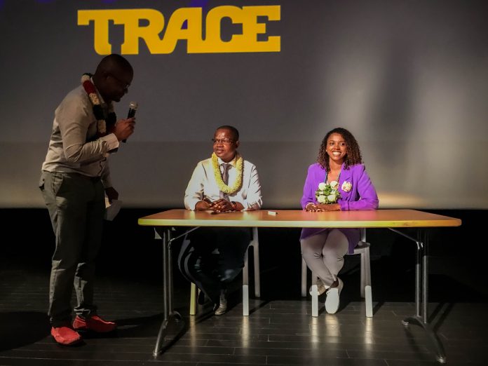 3 personnes dont 2 assises dans une salle sombre, certainement un meeting